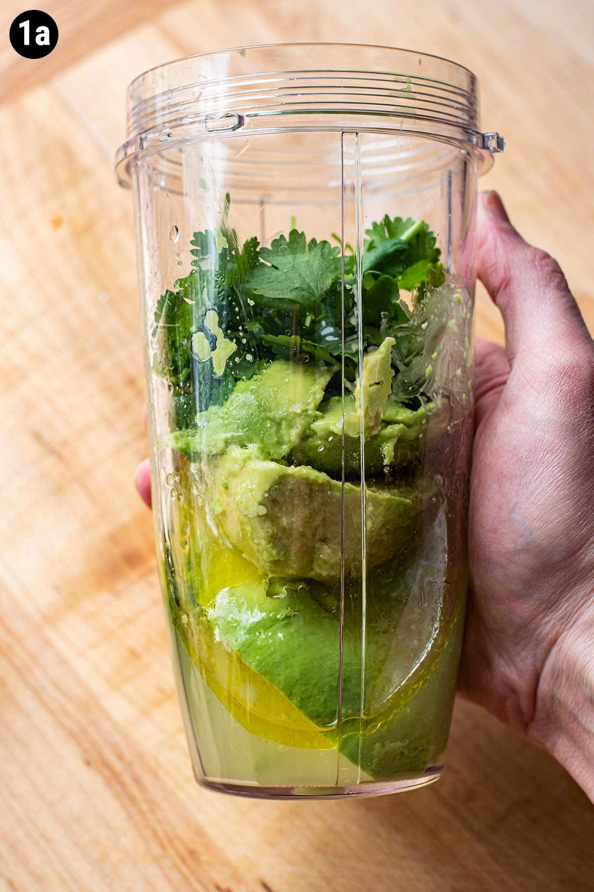A hand holding a blender bottle filled with unblended avocado, fresh cilantro, lime juice, oil, and water.