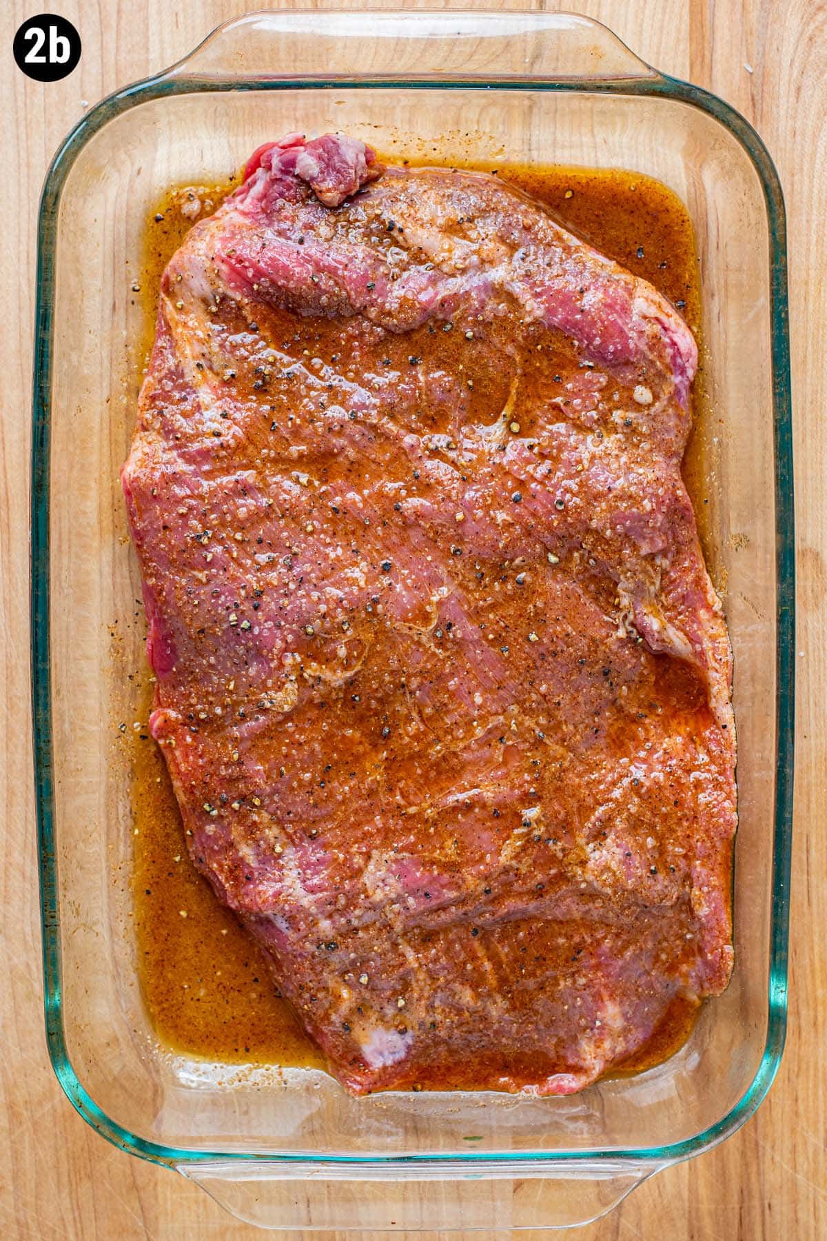 Raw, uncooked marinating flank steak with spices and oil in a clear baking dish.
