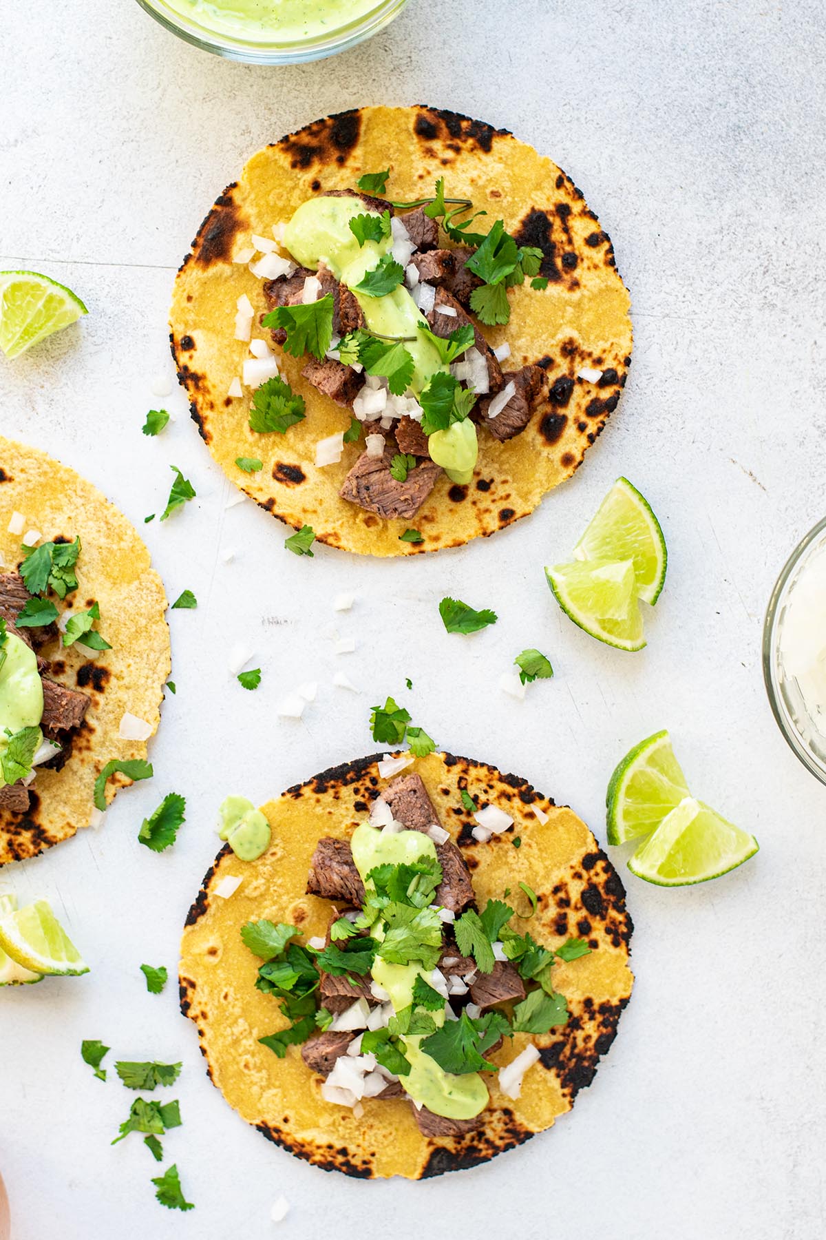 Carne asada tacos on a white table topped with fresh cilantro, chopped white onion, and avocado salsa.