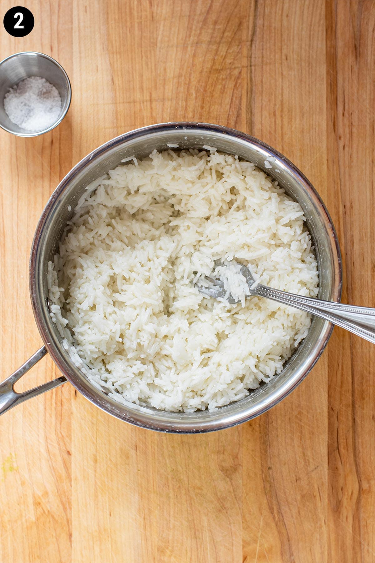 A fork fluffing cooked white rice in a saucepan on a wooden cutting board.
