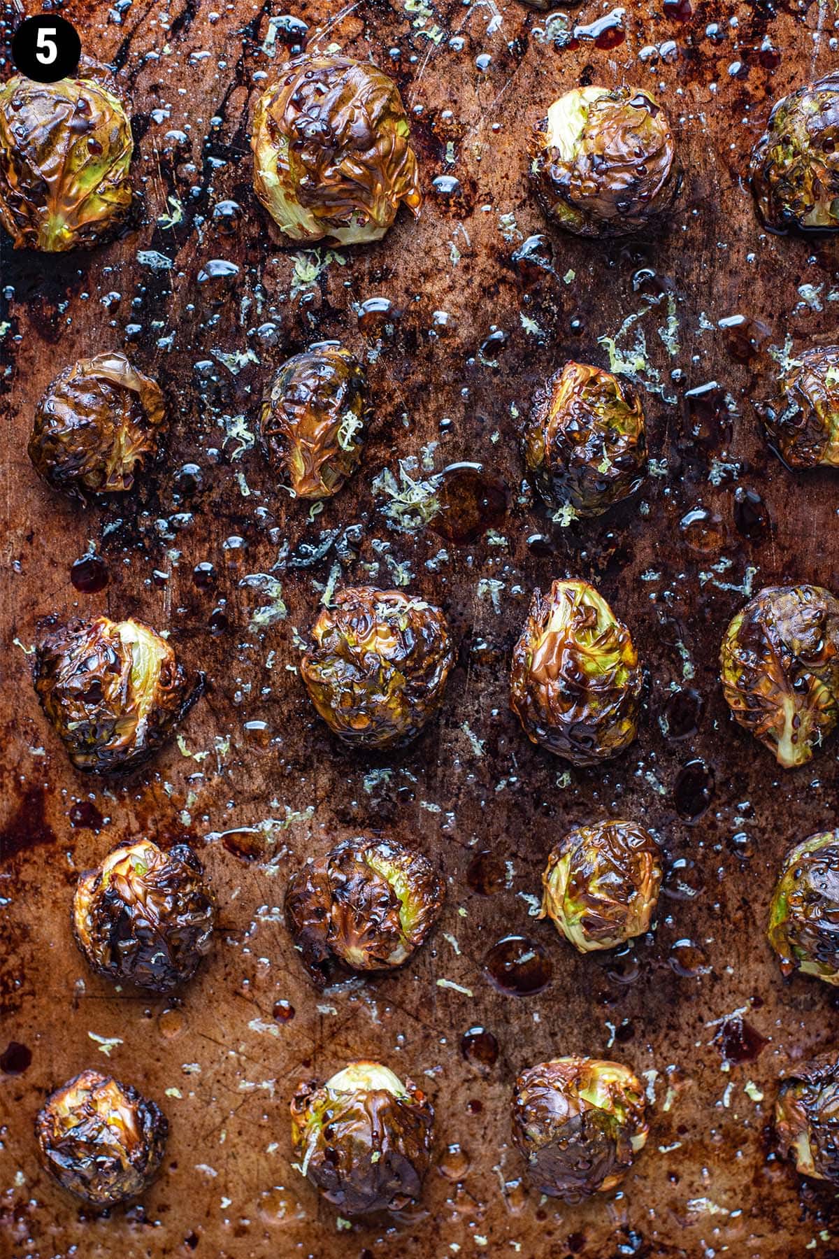 Golden roasted brussels sprouts on a baking sheet topped with balsamic glaze, honey, and lemon zest.