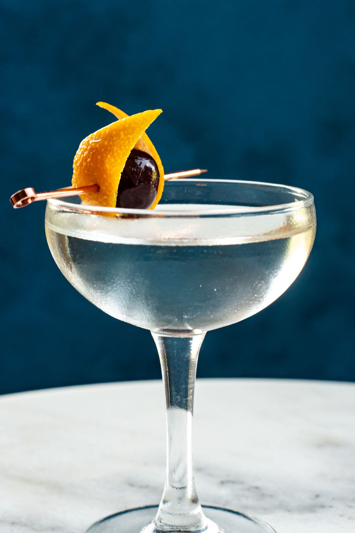 A gin martini garnished with a orange peel and a cherry in a small coupe glass on a marble table.