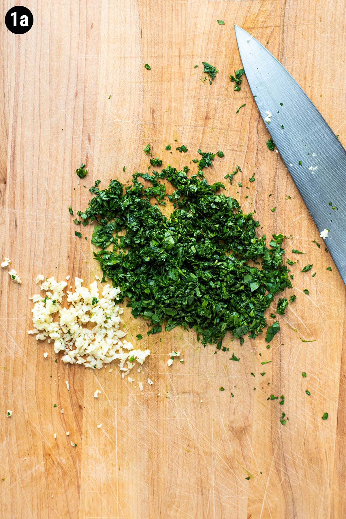 Minced garlic, parsley, and cilantro on a wooden cutting board.
