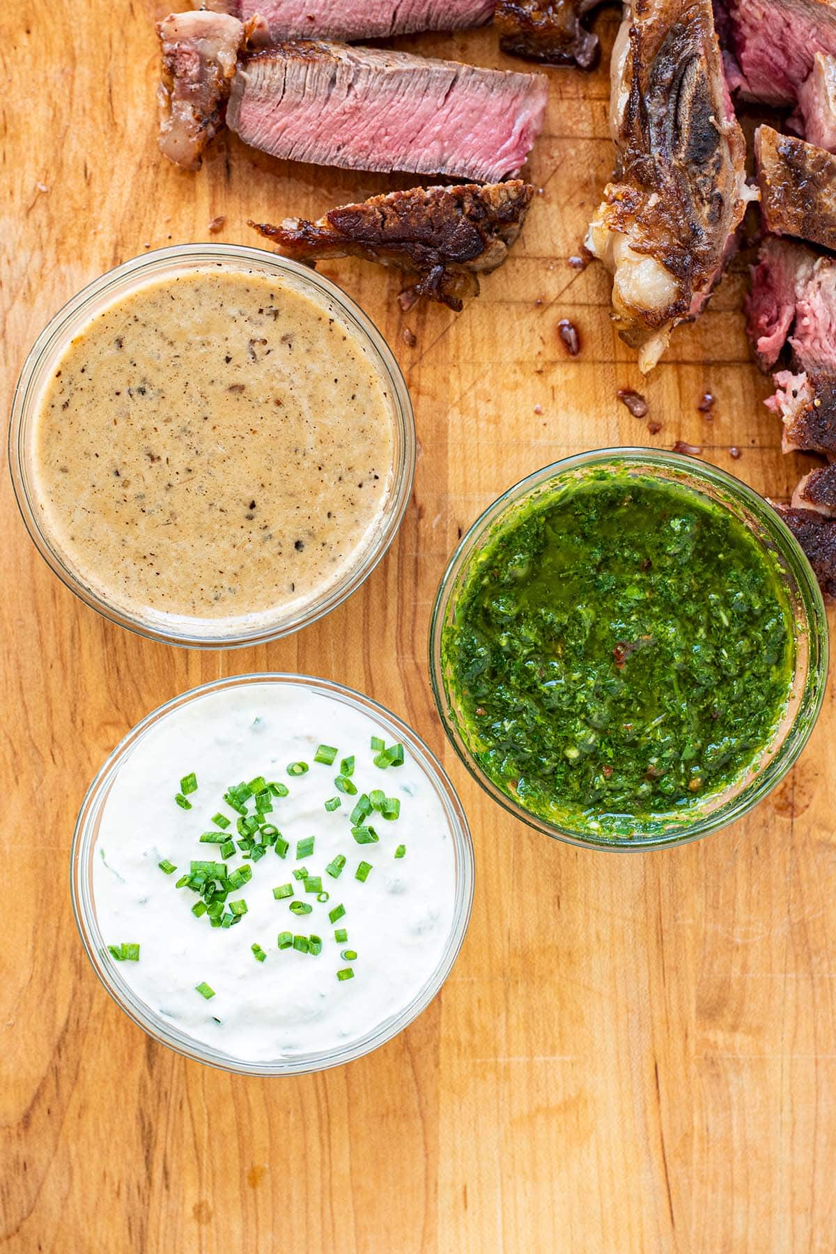 Sliced pink porterhouse steak served with au poivre sauce, chimichurri, and creamy horseradish sauce on a wooden cutting board.