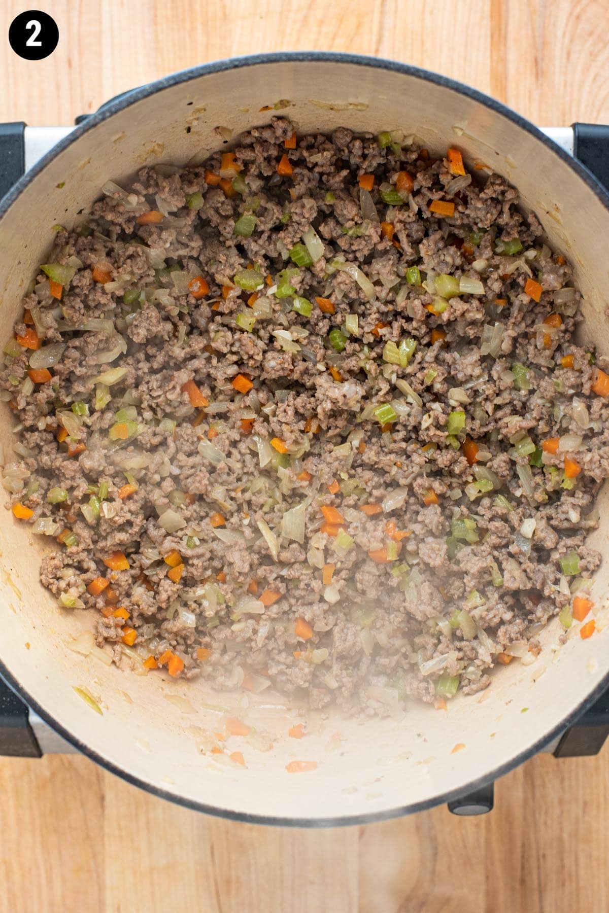 Browned ground beef and sausage cooking in a Dutch oven with chopped carrots, celery, and onion.