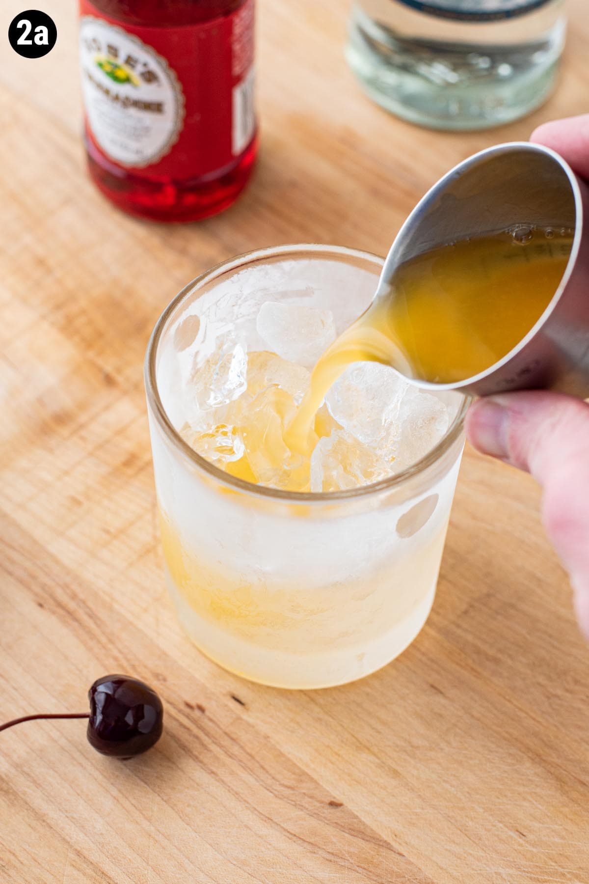 A hand holding a jigger pouring fresh orange juice into an ice filled double old fashioned glass.