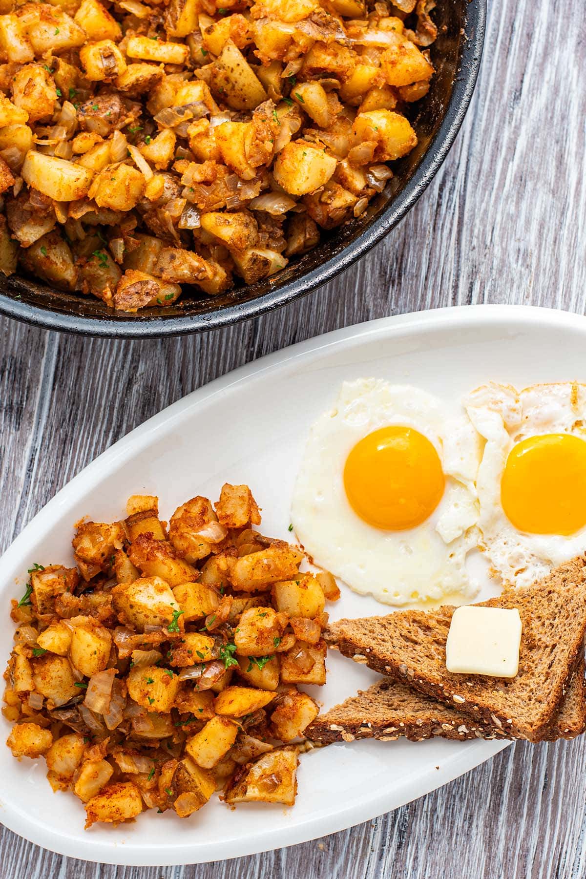 Home fries in a cast iron skillet on a white wooden table next to a plate of home fries, toast, and 2 eggs over easy.