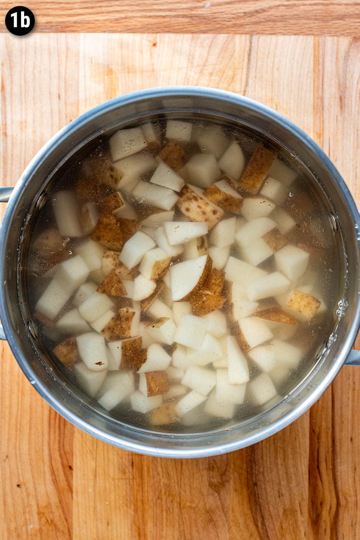 Chopped russet potatoes in a pot full of cold water on a wooden cutting board.