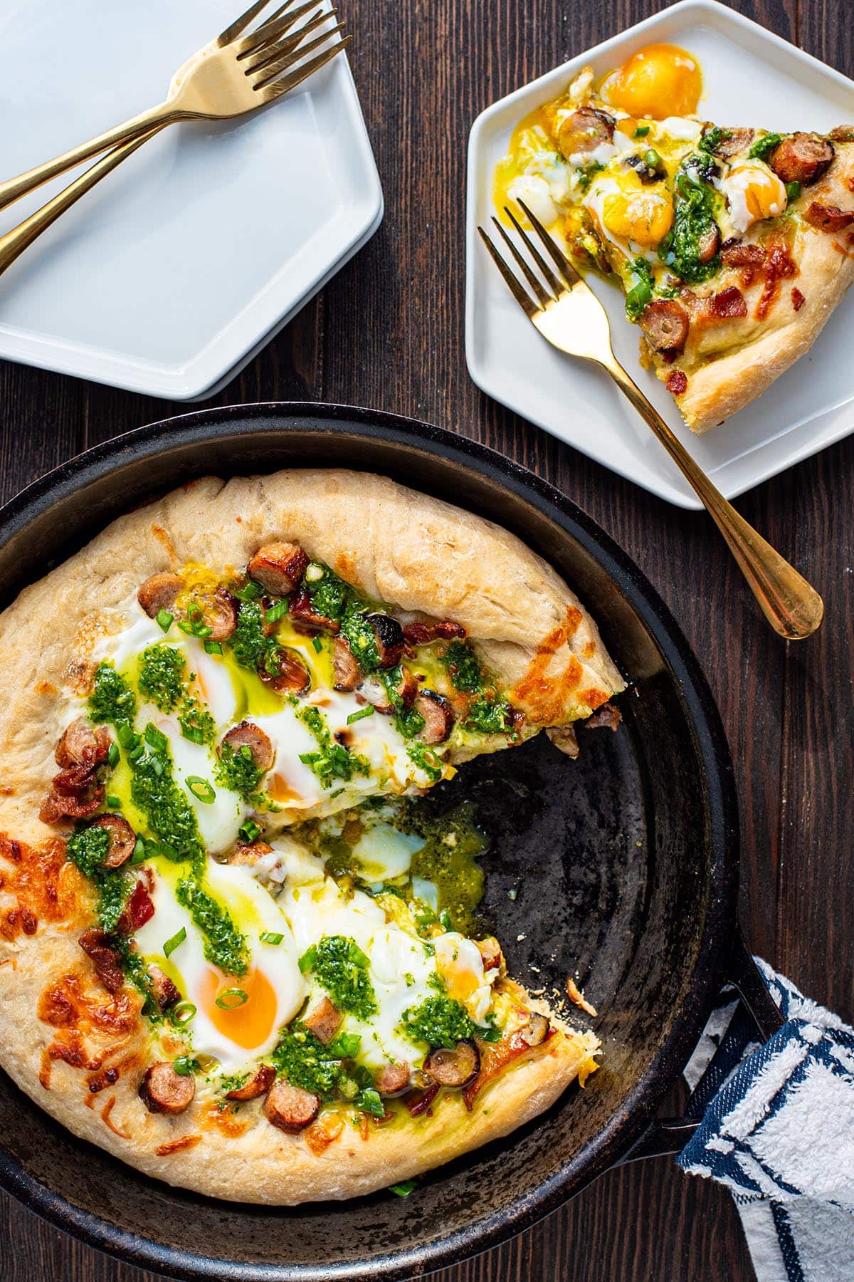 A breakfast pizza in a cast iron skillet with a slice removed, topped with bacon, sausage, runny eggs, and arugula pesto.