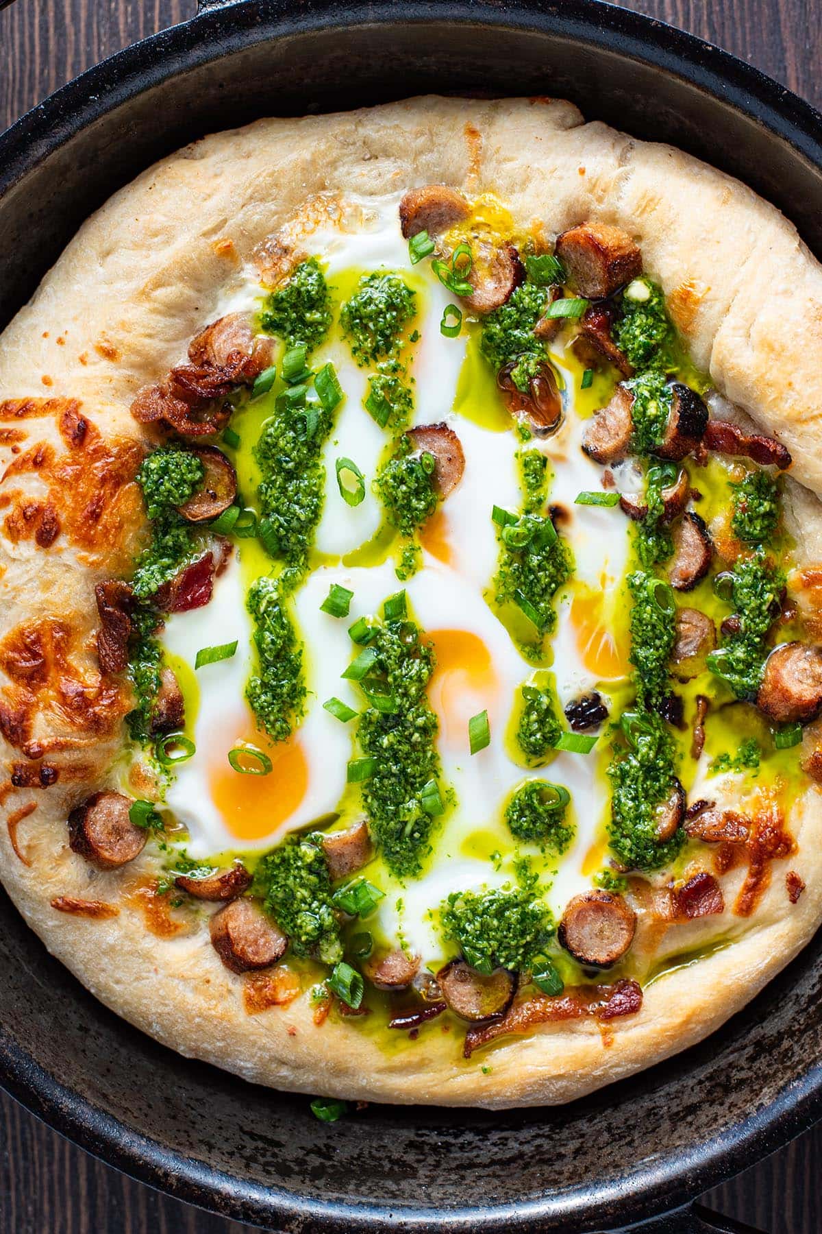 A breakfast pizza in a cast iron skillet topped with bacon, sausage, runny eggs, and arugula pesto.