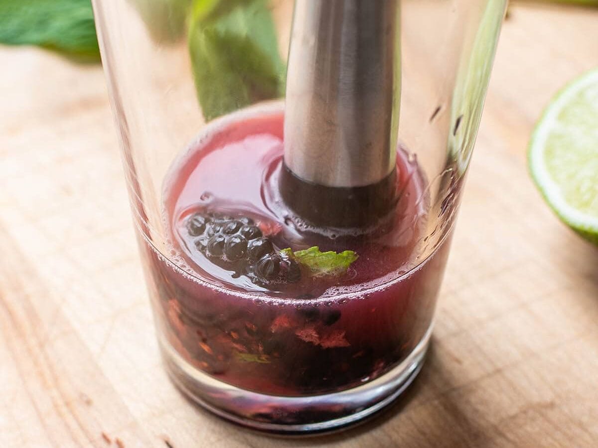 A muddler muddling blackberries and mint leaves in a glass cocktail shaker.
