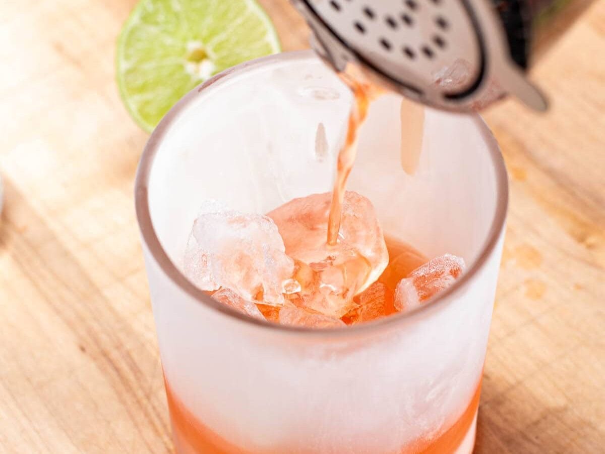Pouring an orange cocktail into a frozen double old fashioned glass with ice.