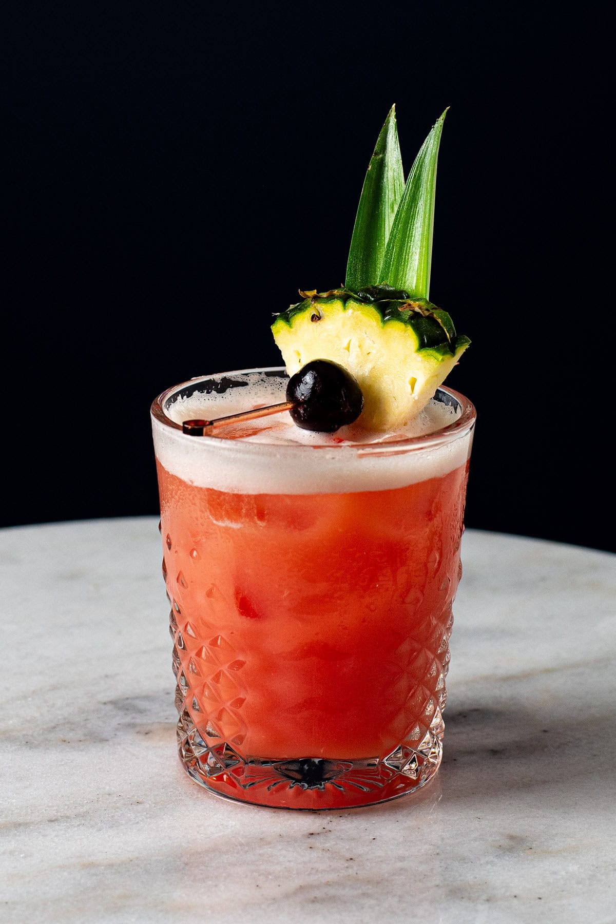 A red-hued Jungle Bird cocktail garnished with a pineapple wedge, pineapple frond, and a brandied cherry.