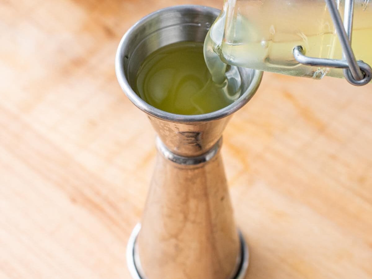 Pouring limoncello into a double jigger on a wooden surface.