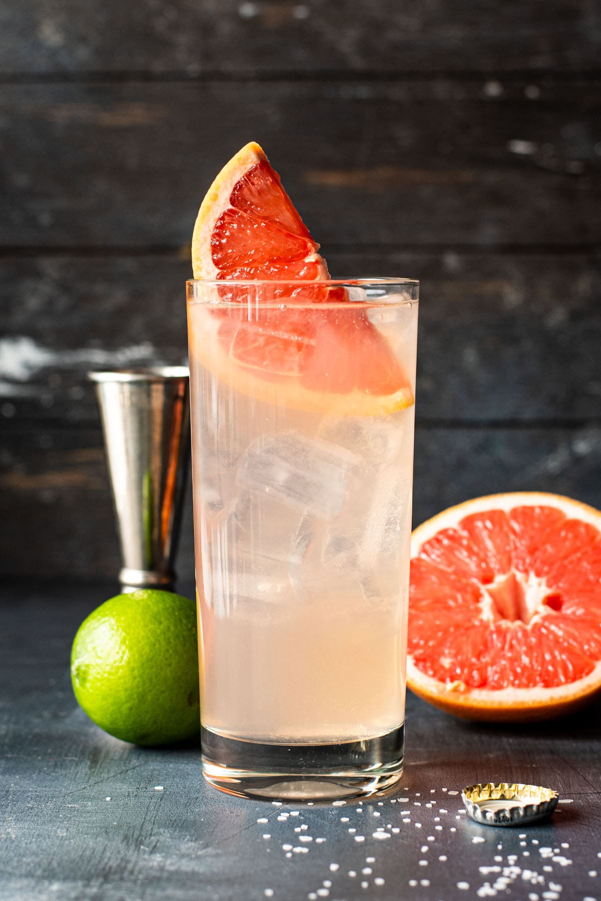 A pink-hued Paloma cocktail garnished with a pink grapefruit wedge on a dark wooden surface next to a lime and sliced grapefruit.
