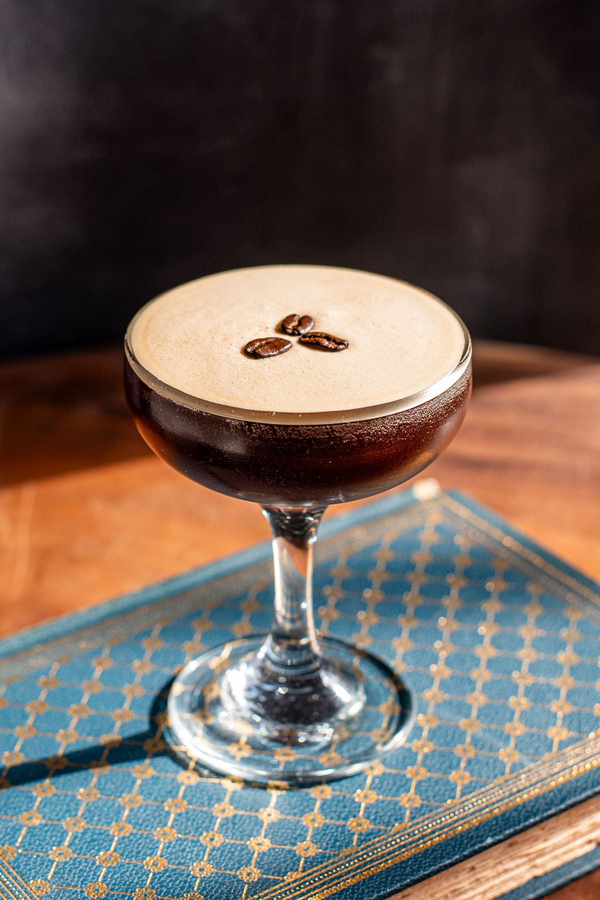 A tequila-based Espresso martini sits on a blue book on a wooden table with a frothy topping and three espresso beans.