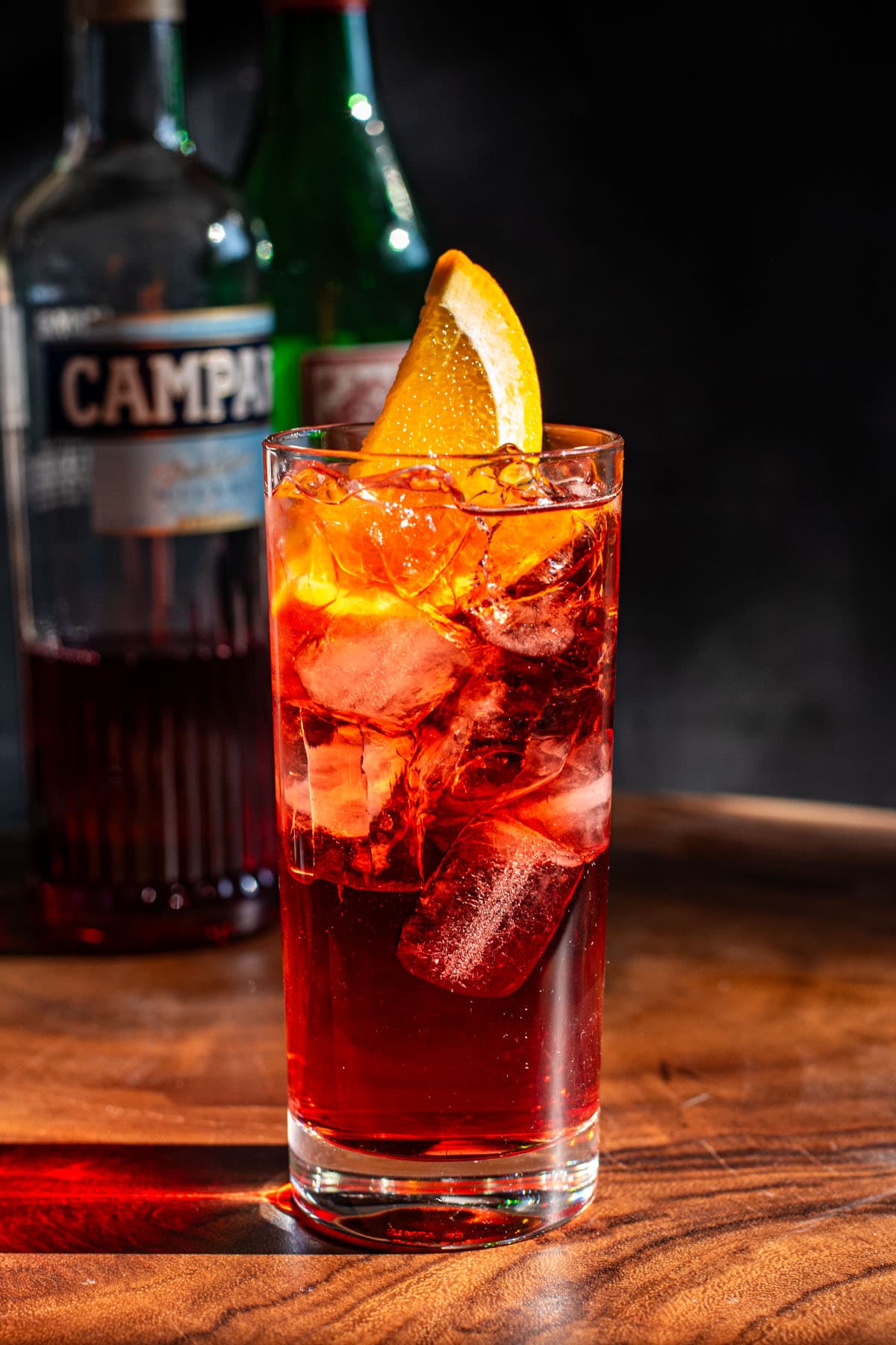 An Americano cocktail on a wooden table in front of a bottle of Campari and sweet vermouth.