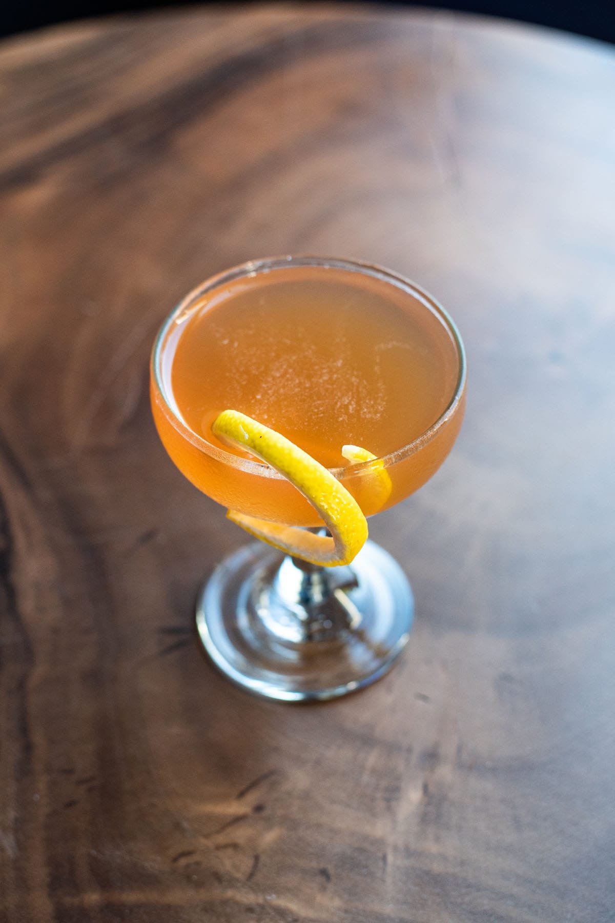An orange hued Champs-Élysées cocktail garnished with a twist of lemon on a wooden table.