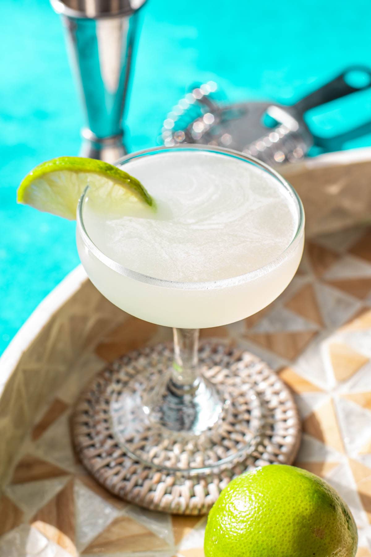 A classic Daiquiri cocktail garnished with a lie wedge on a wooden tray on a bright aqua backdrop.