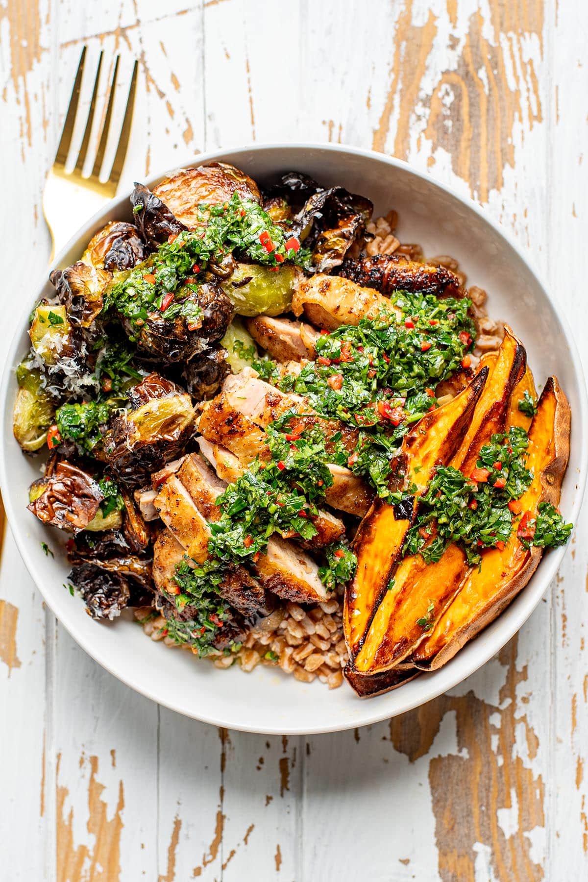 Roasted chicken thighs in a bowl over farro with roasted sweet potato wedges and brussels sprouts topped with fresh herb dressing.