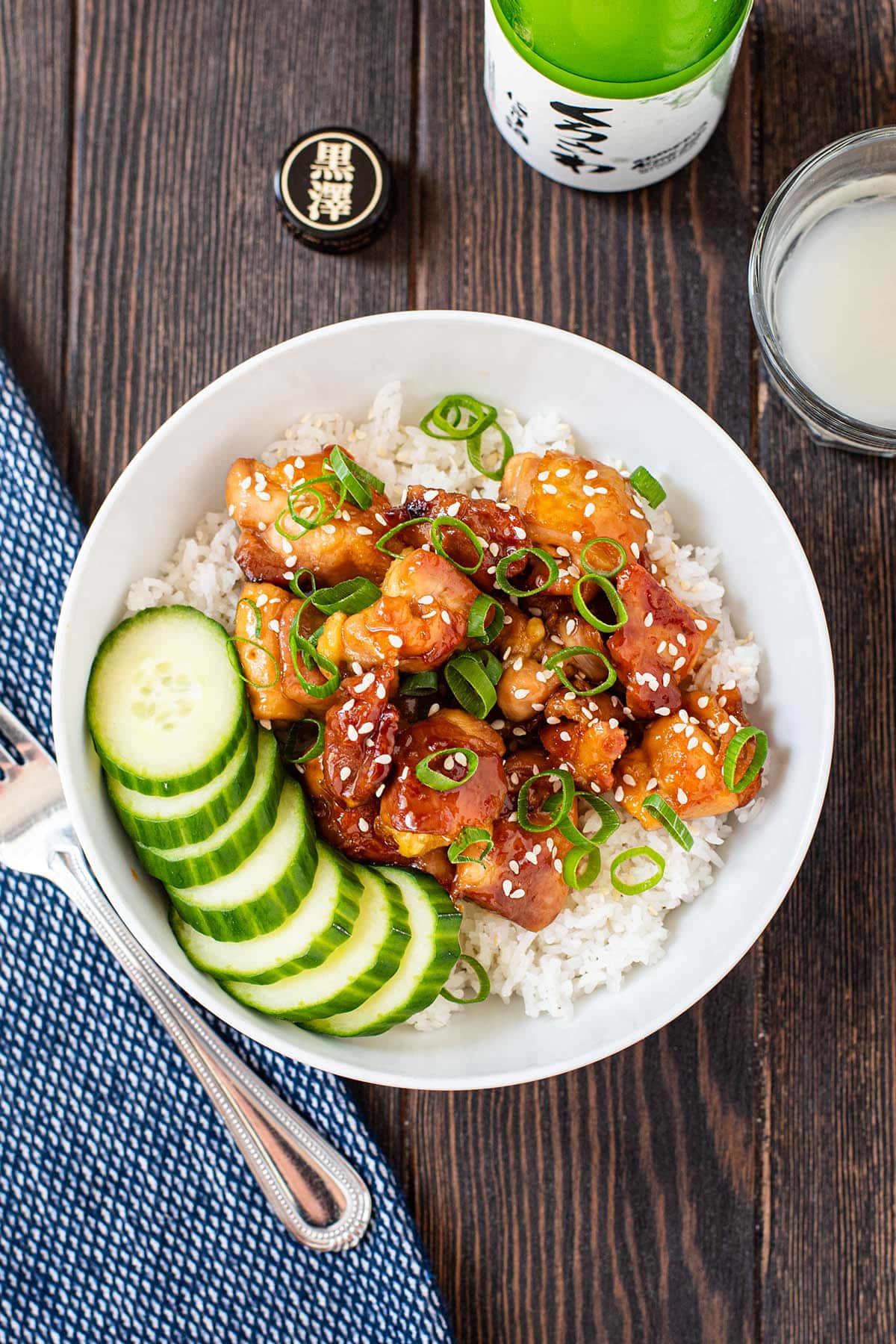 A teriyaki chicken bowl served over white rice and garnished with sliced green onion and sliced cucumber.